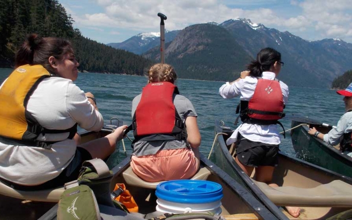four outward bound students sit at the front of canoes. there are mountains in the background. 